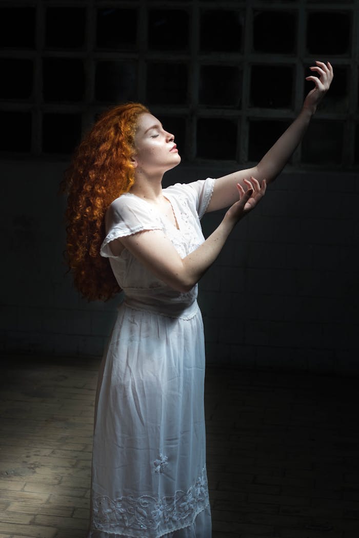 A young red-haired woman in a white dress posed gracefully with dramatic lighting creating an artistic portrait.